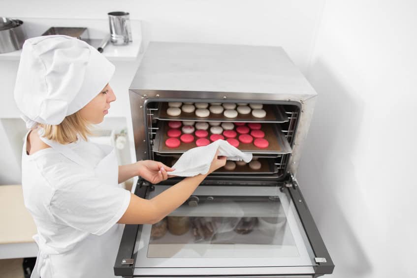 confectionery shop, macarons baking. pretty woman confectioner in white uniform and hat, holds a batch of macarons on baking tray, ready to take it out of the oven after baking.