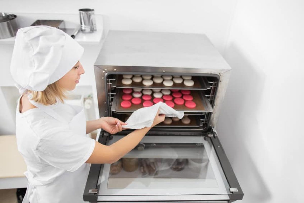 confectionery shop, macarons baking. pretty woman confectioner in white uniform and hat, holds a batch of macarons on baking tray, ready to take it out of the oven after baking.
